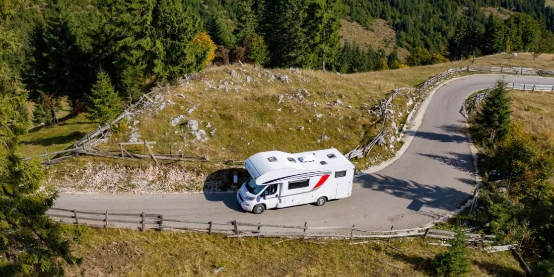 Wei&szlig;es Wohnmobil mit rotem Streifen auf einer kurvigen Bergstra&szlig;e, umgeben von Grash&auml;ngen, Felsen, Holzgel&auml;nder und Nadelb&auml;umen, Luftaufnahme