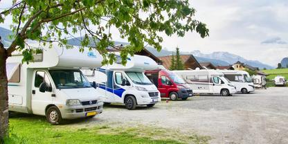 Mehrere Wohnmobile in einer Reihe auf einem Schotter- und Wiesenparkplatz; ein Baum im Vordergrund; H&auml;user und Berge im Hintergrund unter einem bew&ouml;lkten Himmel