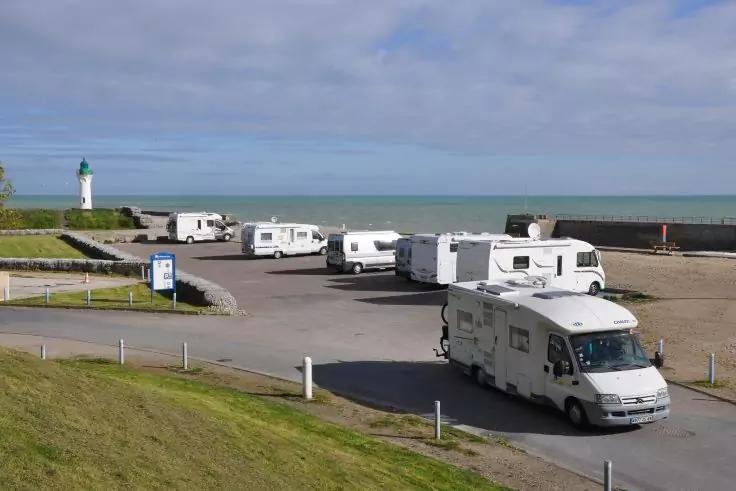 Parkplatz am Meer mit mehreren Wohnmobilen, Leuchtturm links, Gras im Vordergrund und wolkigem Himmel