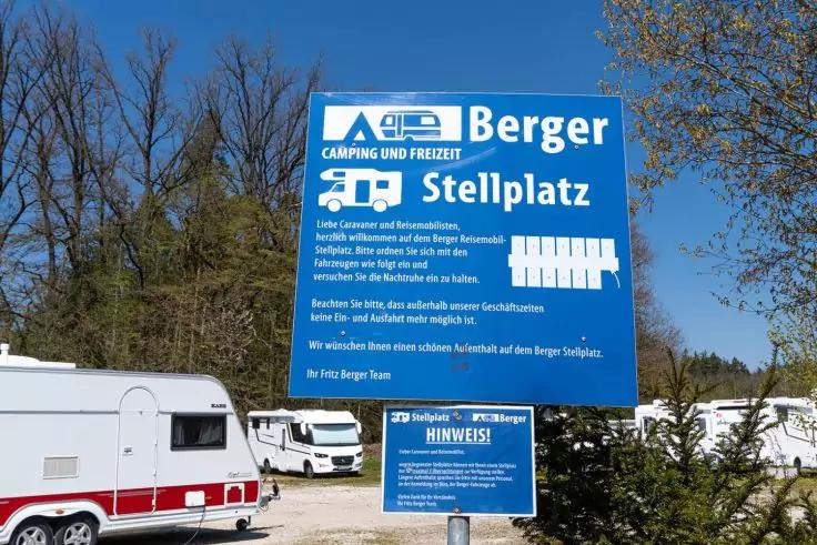 Gro&szlig;es blaues Schild mit wei&szlig;er Aufschrift 'Berger Camping und Freizeit Stellplatz' und Piktogrammen f&uuml;r Wohnmobil und Stellplatz, davor mehrere wei&szlig;e Wohnwagen und Wohnmobile, B&auml;ume ohne Bl&auml;tter und blauer Himmel