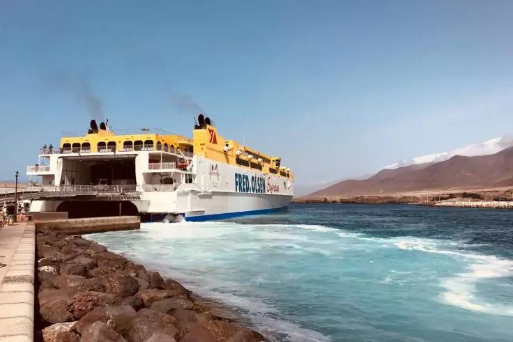Wei&szlig;-gelbes F&auml;hrschiff am Pier, aufgew&uuml;hltes blaues Wasser, Felsenmole links, im Hintergrund schneebedeckte Berge und klarer Himmel