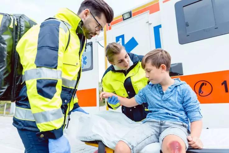 Zwei Rettungskr&auml;fte in gelben Warnjacken, eine beugt sich &uuml;ber einen Jungen mit aufgesch&uuml;rften, blutigen Knien, der auf einer Trage vor einem Krankenwagen sitzt