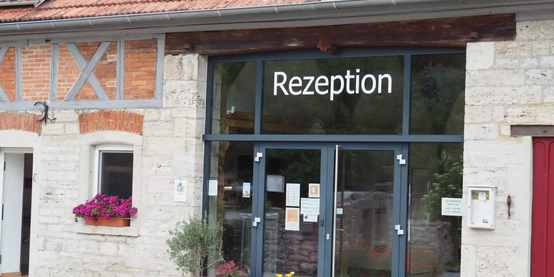 Eingang mit Glast&uuml;r und Schild "Rezeption" &uuml;ber der T&uuml;r, Steinfassade, Fenster mit Blumenkasten, Aush&auml;nge an der T&uuml;r