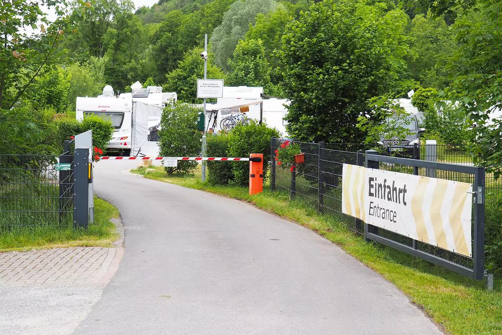 Einfahrt mit Schranke quer &uuml;ber dem Fahrweg, gepflasterte Zufahrt zu mehreren wei&szlig;en Wohnmobilen, Schild "Einfahrt/Entrance", B&auml;ume und Zaun