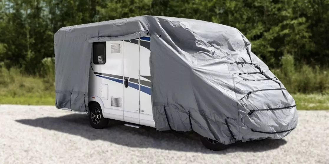 Wei&szlig;es Wohnmobil mit blauem Streifen, teilweise mit grauer Abdeckung &uuml;ber Front und Dach, steht auf Kies vor B&auml;umen