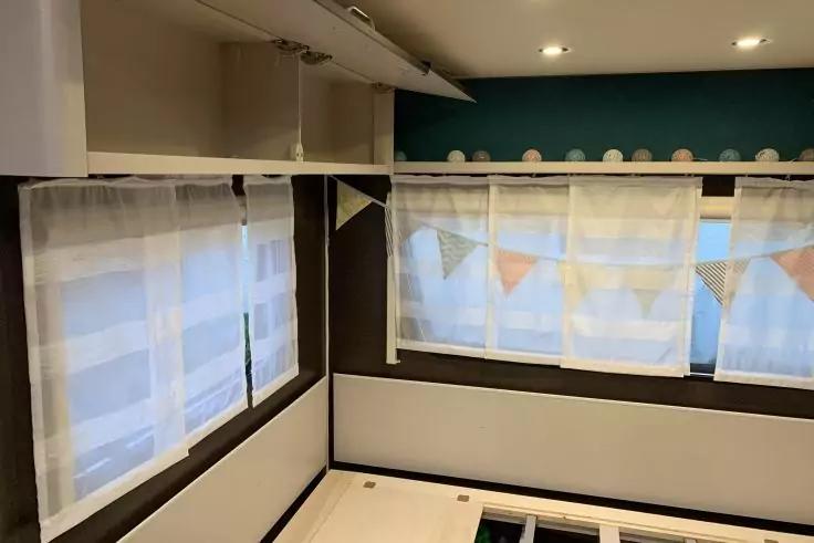 Drei Fenster mit wei&szlig;en Gardinen; Wimpelkette &uuml;ber dem mittleren Fenster; Regal mit kleinen runden Gegenst&auml;nden; dunkle und helle Wandverkleidung; drei Deckenleuchten; helles M&ouml;belgestell mit Latten