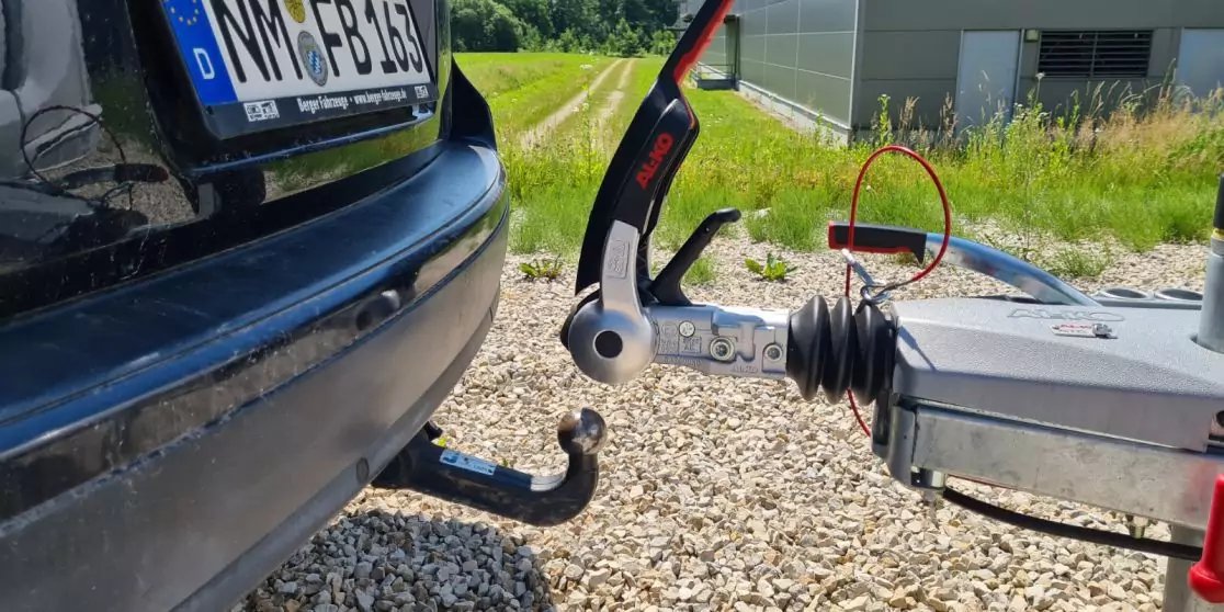 Heck eines schwarzen Autos mit Kugel-Anh&auml;ngerkupplung, an eine Anh&auml;ngerzugvorrichtung gekoppelt; Schotterboden, Gras und Geb&auml;ude im Hintergrund