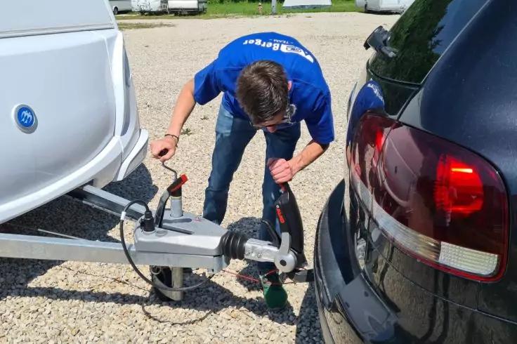 Person beugt sich zwischen dem Heck eines schwarzen Autos und einem Anh&auml;nger und befestigt die Anh&auml;ngerkupplung