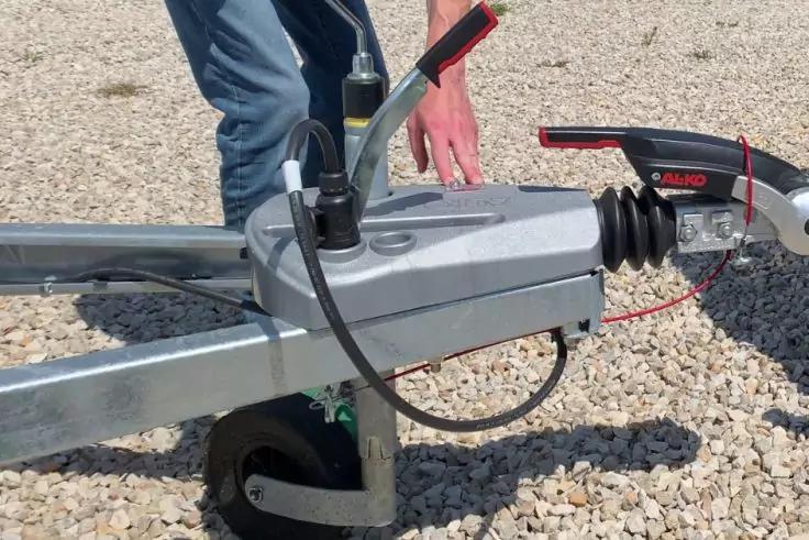 Hand an Anh&auml;ngerkupplung mit St&uuml;tzrad und Kabeln auf Kiesboden