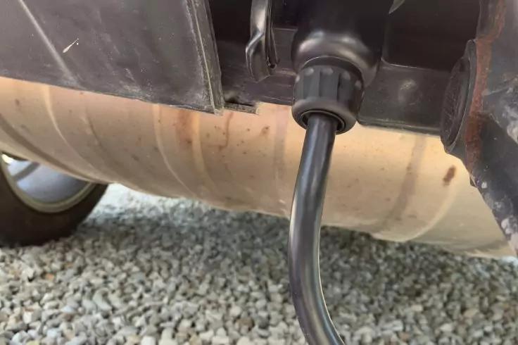 Unterseite eines Fahrzeugs mit Auspuffschalld&auml;mpfer, daran ein schwarzes Kabel mit Steckverbindung, Kiesboden darunter