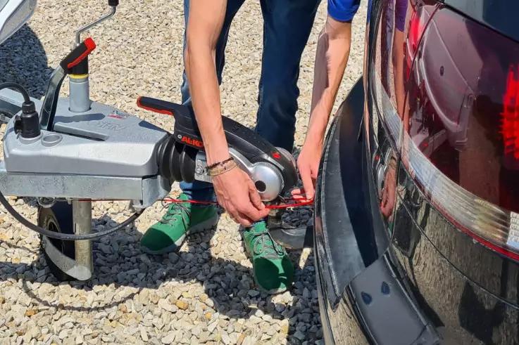 Arme und Beine einer Person, die auf Kies eine Anh&auml;ngerkupplung an der R&uuml;ckseite eines dunklen Autos befestigt