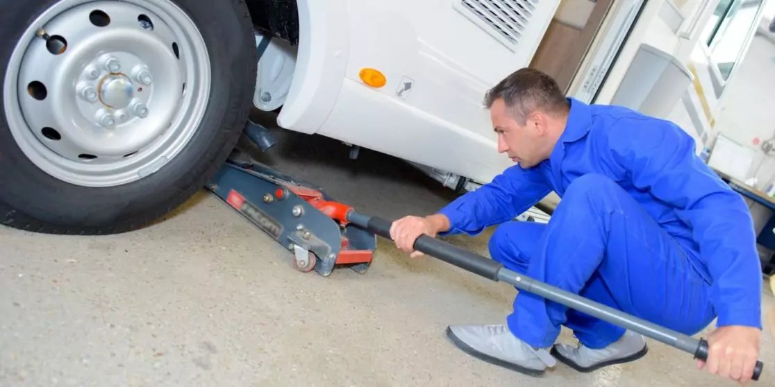 Person in blauer Arbeitskleidung kniet neben einem wei&szlig;en Fahrzeug und bedient einen Wagenheber unter dem Fahrzeug nahe dem Rad