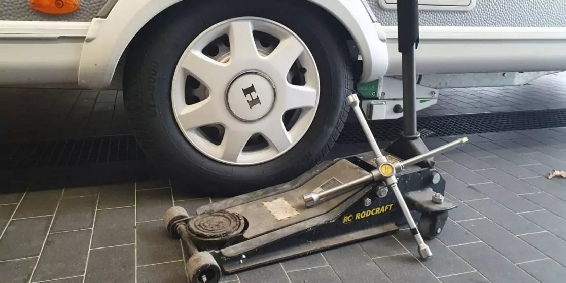 Nahaufnahme eines wei&szlig;en Wohnwagenrads mit Kunststoffradkappe, darunter ein hydraulischer Wagenheber auf grauem Fliesenboden