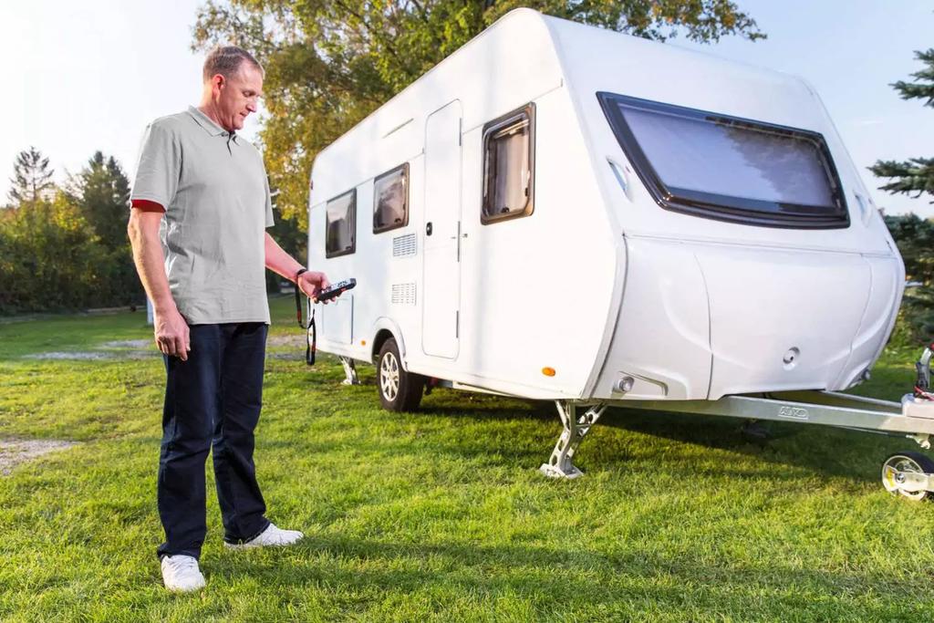 Ein Mann steht auf einer Wiese neben einem wei&szlig;en Wohnwagen und h&auml;lt eine Schl&uuml;ssel- oder Fernbedienung; B&auml;ume im Hintergrund