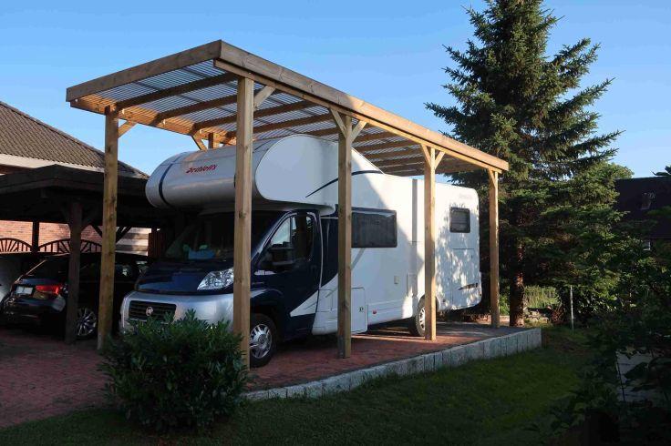 Wei&szlig;es Wohnmobil unter einem h&ouml;lzernen Carport mit transluzentem Dach, neben einem hohen Nadelbaum; Rasen und Strauch im Vordergrund, H&auml;user und ein weiteres geparktes Auto im Hintergrund