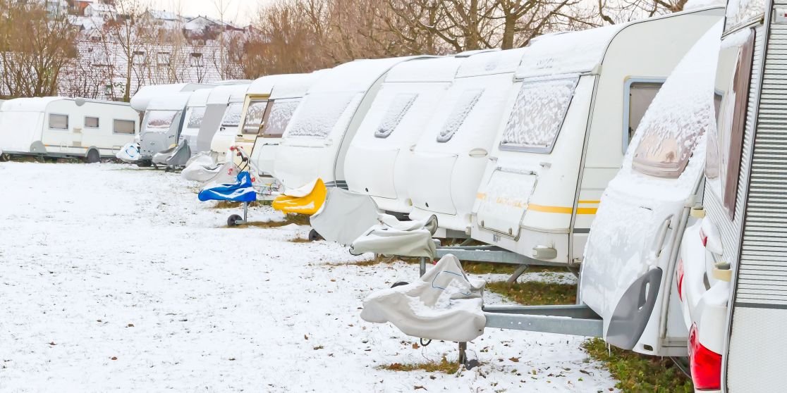 Reihe wei&szlig;er Wohnwagen auf schneebedecktem Boden; vor einigen Wagen kleine Boote auf Anh&auml;ngern; B&auml;ume und Geb&auml;ude im Hintergrund
