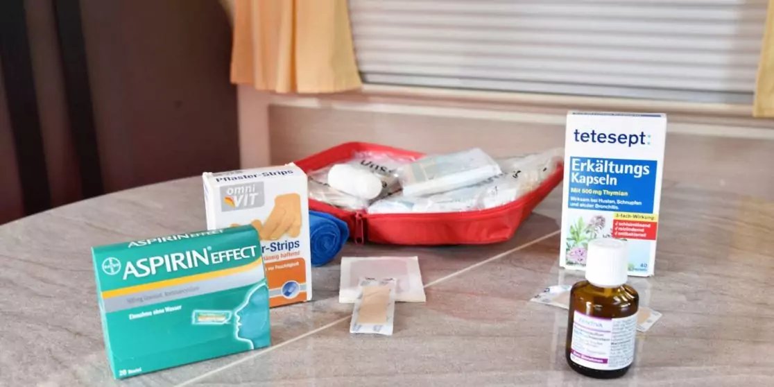 Tisch mit mehreren Medikamentenverpackungen: gr&uuml;ne Aspirin-Schachtel, kleine Tetesept-Packung, braune Flasche mit Etikett, rotes Kunststoffsch&auml;lchen mit wei&szlig;en T&uuml;chern, zwei einzelne Verpackungen auf dem Tisch; im Hintergrund Fenster mit geschlossene