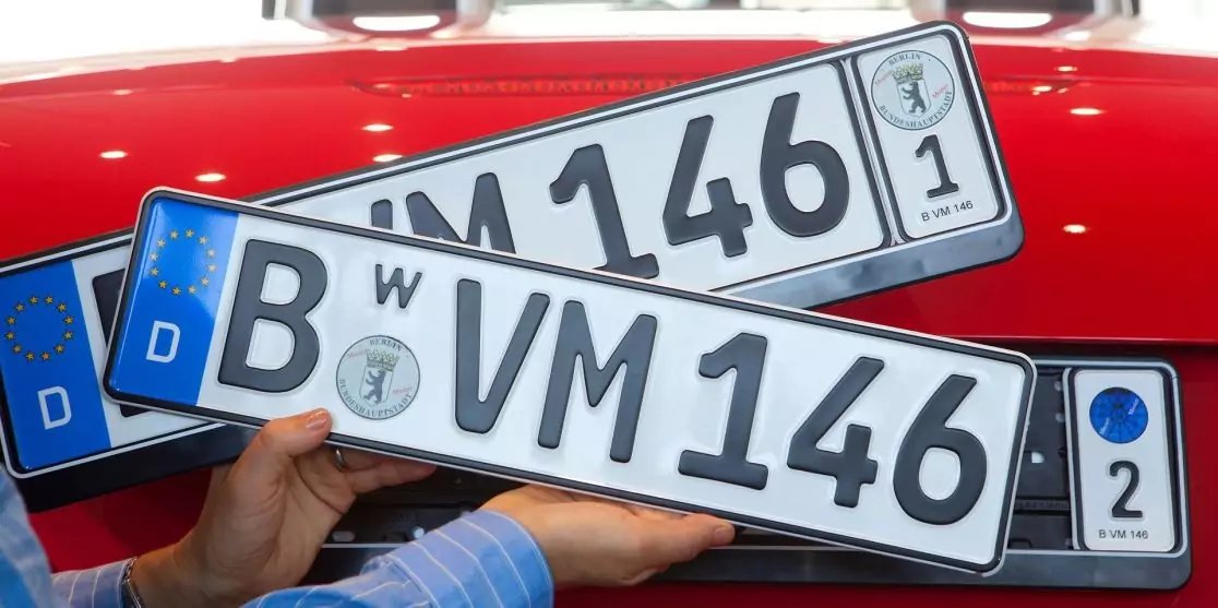 Two German license plates ('B VM 146' and 'W VM 146') stacked on a red car hood, hands holding the front plate