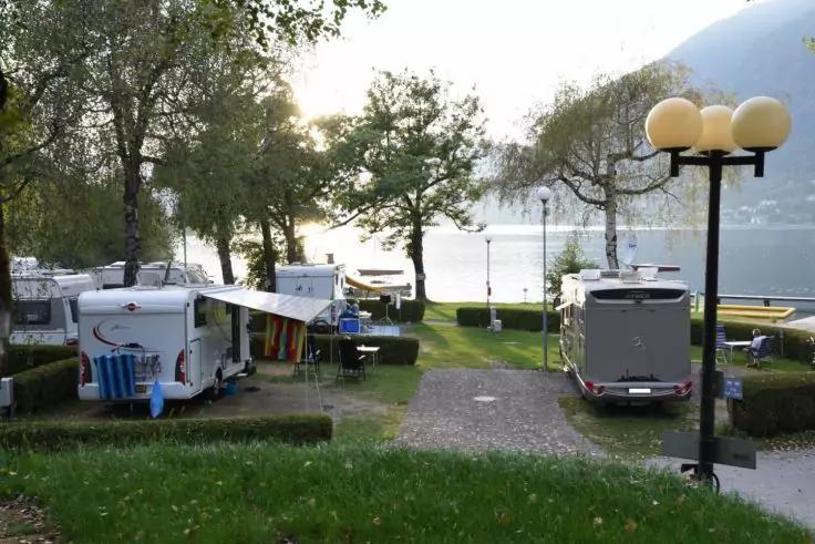 Zwei Wohnwagen mit aufgeklappter Markise neben einem grasbewachsenen Weg zur Uferlinie, Laternenmast, B&auml;ume, Sitzb&auml;nke, Boote am Wasser, See mit Bergen im Hintergrund