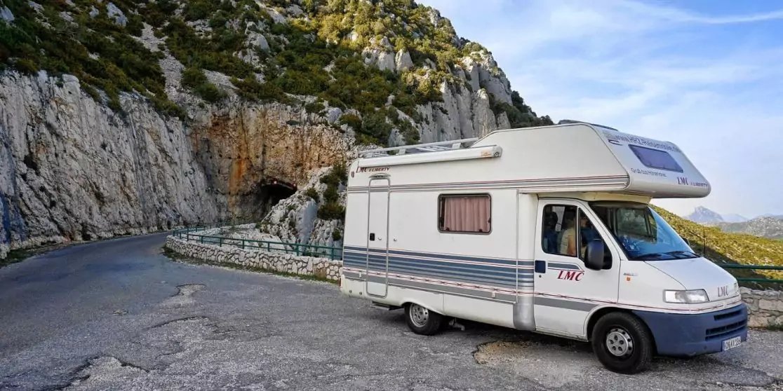 Wei&szlig;es Wohnmobil auf einer Bergstra&szlig;e vor einem Felstunnel, steinerne Leitplanke und blauer Himmel