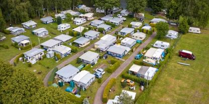 Luftaufnahme mehrerer Wohnwagen und kleiner Mobilheime in Reihen auf einer Gr&uuml;nfl&auml;che mit Wegen und B&auml;umen, roter Van am Rand