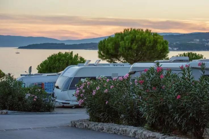 Mehrere Wohnwagen hinter bl&uuml;henden Str&auml;uchern an der K&uuml;ste, im Hintergrund Meer, H&uuml;gel und ein Baum unter rosa-gelbem Himmel