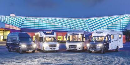 F&uuml;nf Wohnmobile und Vans nebeneinander auf gepflastertem Platz vor einem Geb&auml;ude mit gro&szlig;em, beleuchtetem Glasdach bei D&auml;mmerung