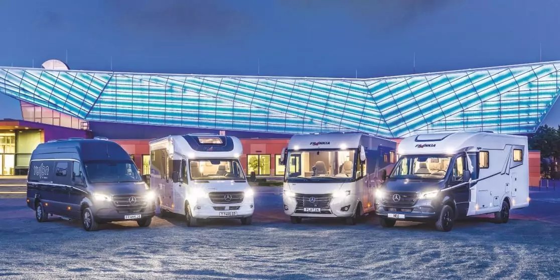 F&uuml;nf Wohnmobile und Vans nebeneinander auf gepflastertem Platz vor einem Geb&auml;ude mit gro&szlig;em, beleuchtetem Glasdach bei D&auml;mmerung