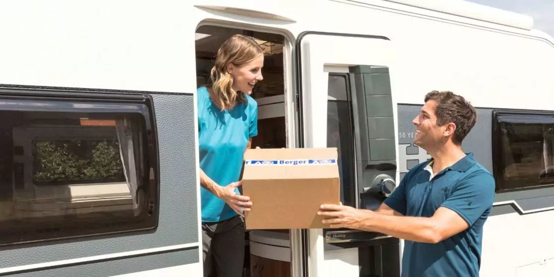 Woman in the open door of a motorhome hands a cardboard box to a man outside