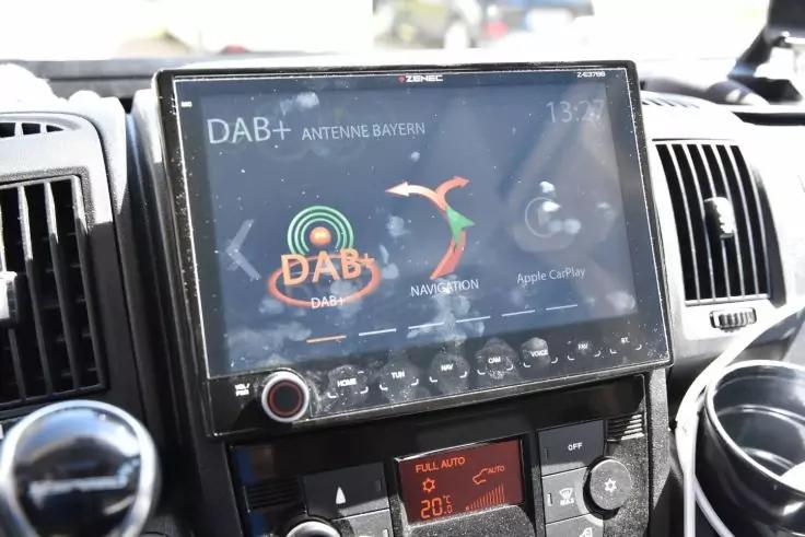 Zentrales Touchscreen im Armaturenbrett zeigt DAB+-Radioanzeige mit Symbolen; links und rechts L&uuml;ftungsd&uuml;sen; darunter Klimabedienelemente und rechts ein Getr&auml;nkehalter mit Becher