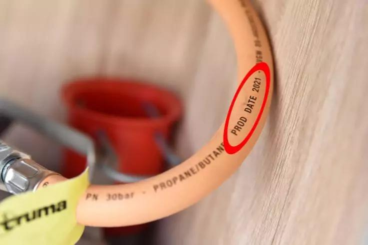 Orangener flexibler Schlauch mit aufgedrucktem Text 'PROD DATE 2021', der Text ist mit einem roten Kreis markiert; Metallanschluss mit gelbem Schutz links; roter Kunststoffbeh&auml;lter im Hintergrund; Holzwand rechts