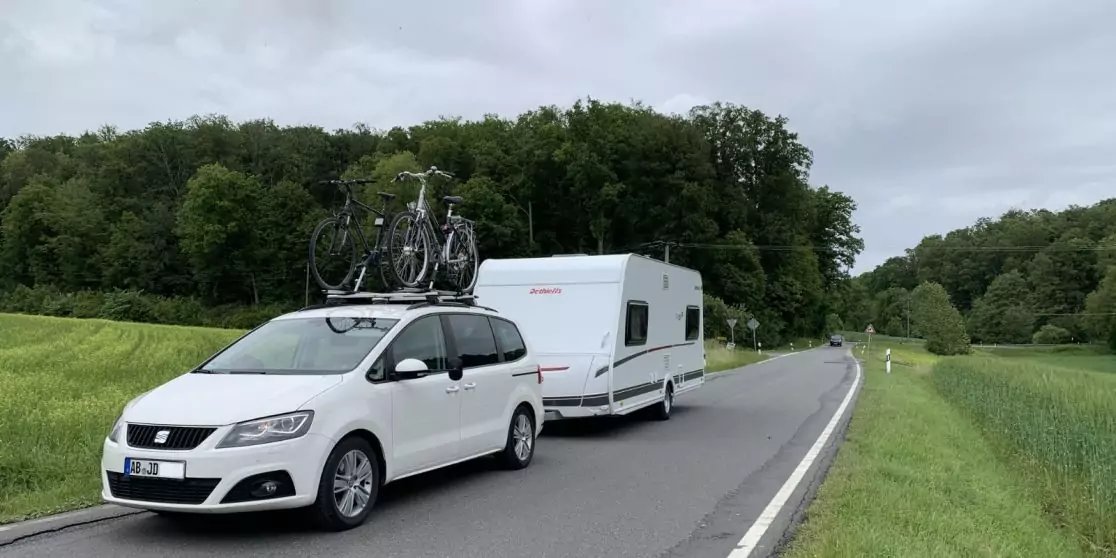 White car with attached caravan on a country road, two bicycles on the car roof, green fields and trees, cloudy sky
