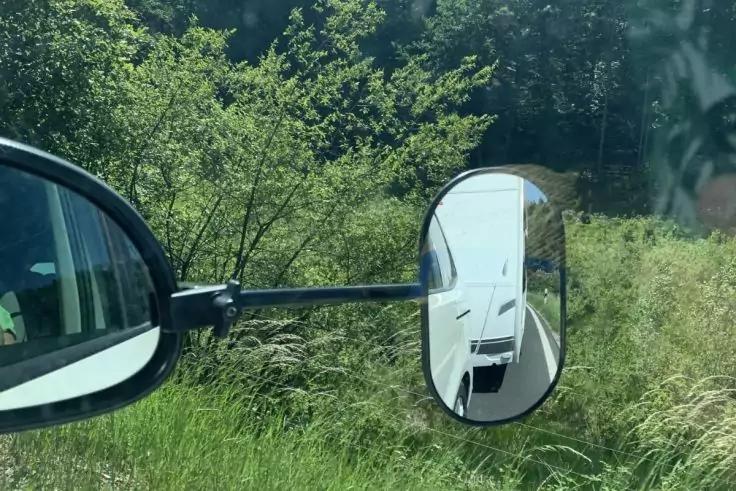 Blick aus einem Auto: Au&szlig;enspiegel und zus&auml;tzlicher Anh&auml;ngerspiegel; im Anh&auml;ngerspiegel ein wei&szlig;er Wohnwagen; Gras und B&auml;ume am Stra&szlig;enrand