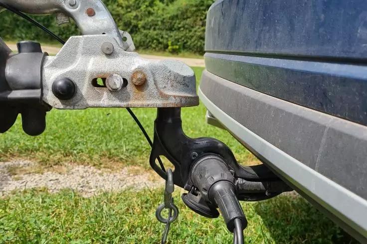 Nahaufnahme einer Anh&auml;ngerkupplung am Heck eines Fahrzeugs mit eingestecktem Elektrostecker, befestigter Sicherheits&ouml;se und Gras sowie Geb&uuml;sch im Hintergrund
