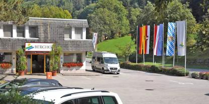 Eingangsbereich eines Geb&auml;udes mit Schild, vorne ein wei&szlig;er Transporter und geparkte Autos, rechts mehrere Flaggen auf Fahnenmasten, im Hintergrund bewaldete H&uuml;gel