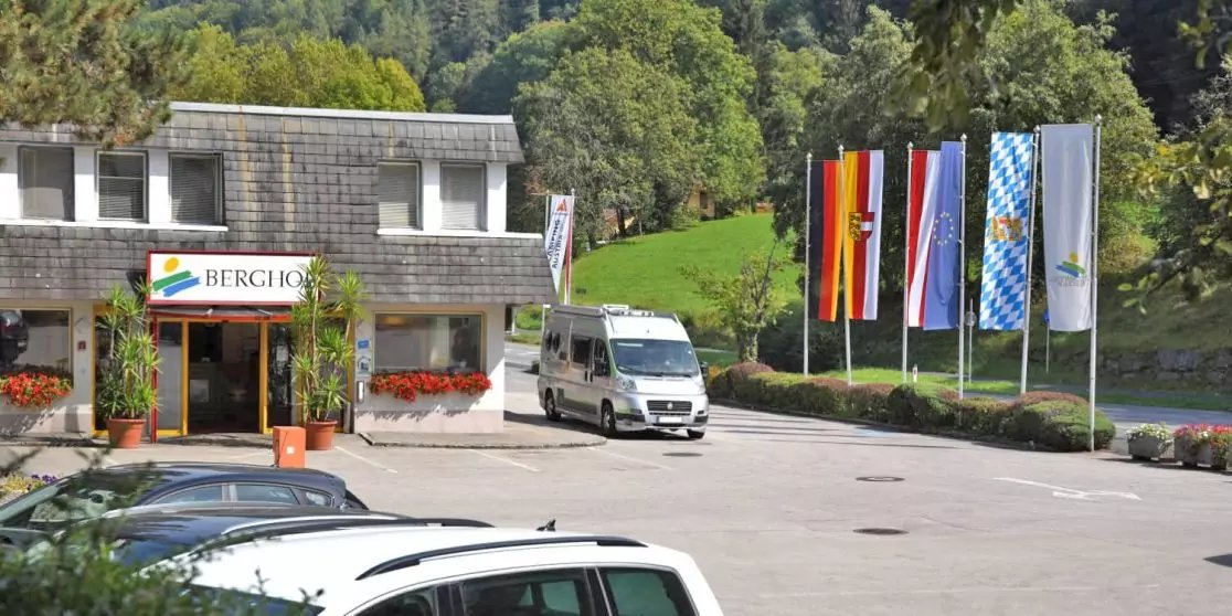 Eingangsbereich eines Geb&auml;udes mit Schild, vorne ein wei&szlig;er Transporter und geparkte Autos, rechts mehrere Flaggen auf Fahnenmasten, im Hintergrund bewaldete H&uuml;gel