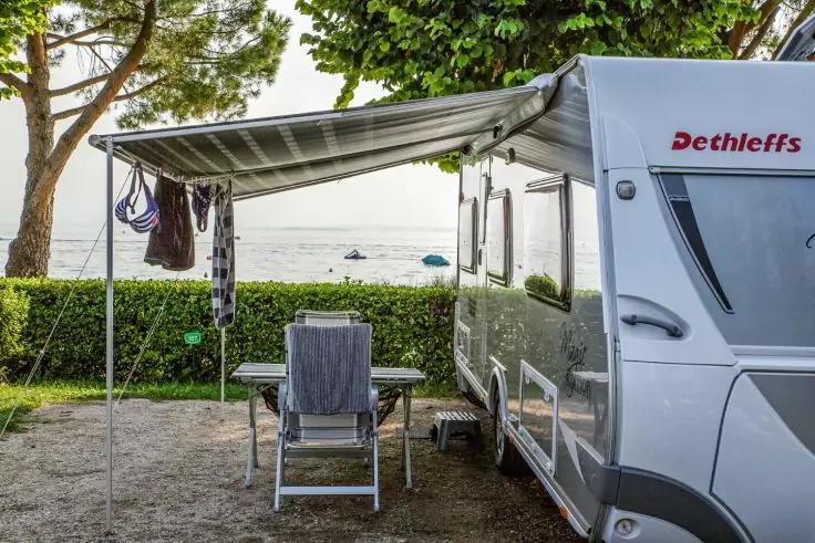 Wei&szlig;er Wohnwagen mit ausgefahrener Markise, Tisch und zwei St&uuml;hlen, Handtuch &uuml;ber einem Stuhl, Hecke und B&auml;ume vor dem Meer mit Booten