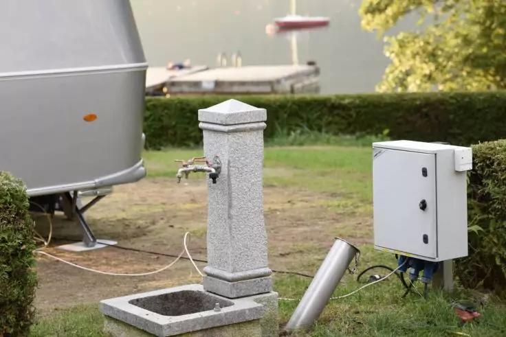 Graue steinerne Wassers&auml;ule mit Metallhahn und rechteckiger Auffangrinne, daneben ein wei&szlig;er Elektrokasten mit sichtbaren Kabeln, ein liegendes Metallrohr, links Teile eines Wohnwagens; im Hintergrund Rasen, Hecke, See mit kleinem Steg und runder Sch