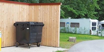 Gro&szlig;er schwarzer M&uuml;llcontainer auf Rollen vor einer Holzsichtschutzwand; links teilweise eine gelbe Tonne, rechts eine Rasenfl&auml;che mit hellem Wohnwagen im Hintergrund, ein Steinblock neben einem gepflasterten Weg und B&auml;ume
