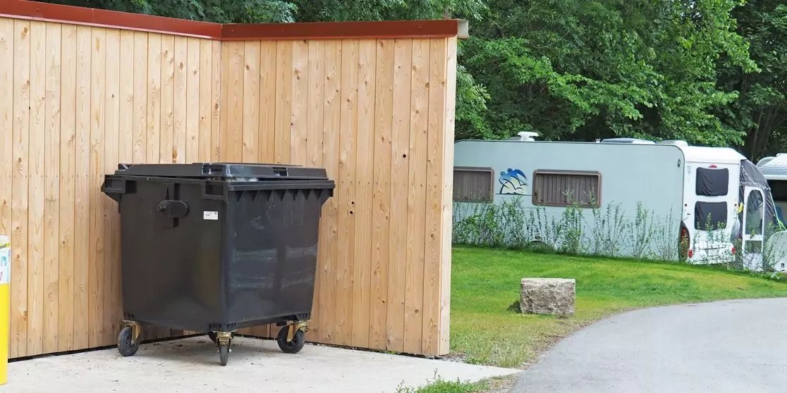 Gro&szlig;er schwarzer M&uuml;llcontainer auf Rollen vor einer Holzsichtschutzwand; links teilweise eine gelbe Tonne, rechts eine Rasenfl&auml;che mit hellem Wohnwagen im Hintergrund, ein Steinblock neben einem gepflasterten Weg und B&auml;ume