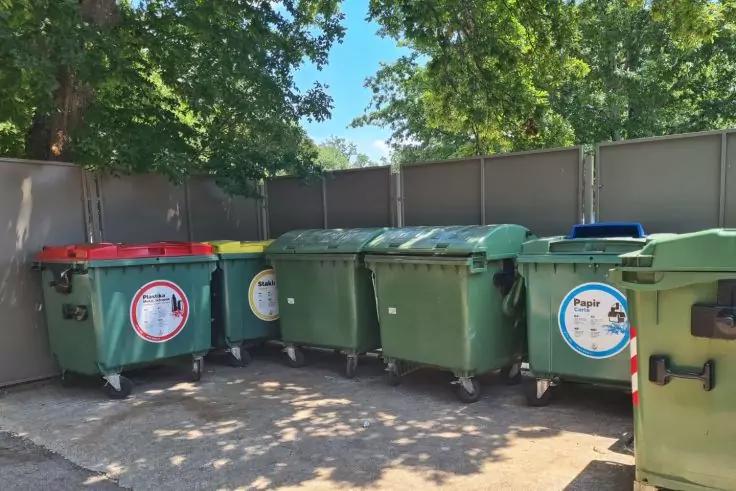 Mehrere gro&szlig;e gr&uuml;ne M&uuml;llcontainer mit farbigen Deckeln und Aufklebern nebeneinander vor einer grauen Umz&auml;unung; B&auml;ume und blauer Himmel im Hintergrund