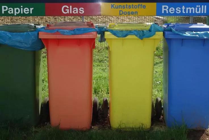 Vier M&uuml;lltonnen (dunkelgr&uuml;n, rot, gelb, blau) nebeneinander auf Gras; dar&uuml;ber farbige Schilder mit den Aufschriften 'Papier', 'Glas', 'Kunststoffe Dosen', 'Restm&uuml;ll'