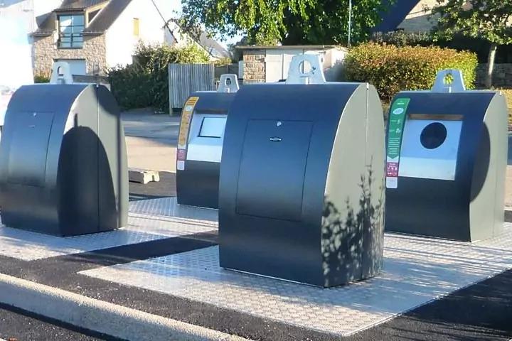 Drei dunkelgraue oberirdische M&uuml;llcontainer mit runden und rechteckigen Einwurf&ouml;ffnungen auf einer gepflasterten Metallplattform; H&auml;user, Hecke und Baumschatten im Hintergrund