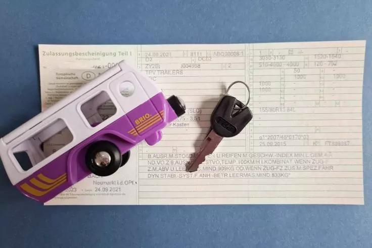 Wei&szlig;-lila Spielzeug-Wohnmobil auf einem gedruckten Formular, daneben ein Autoschl&uuml;ssel auf blauem Untergrund