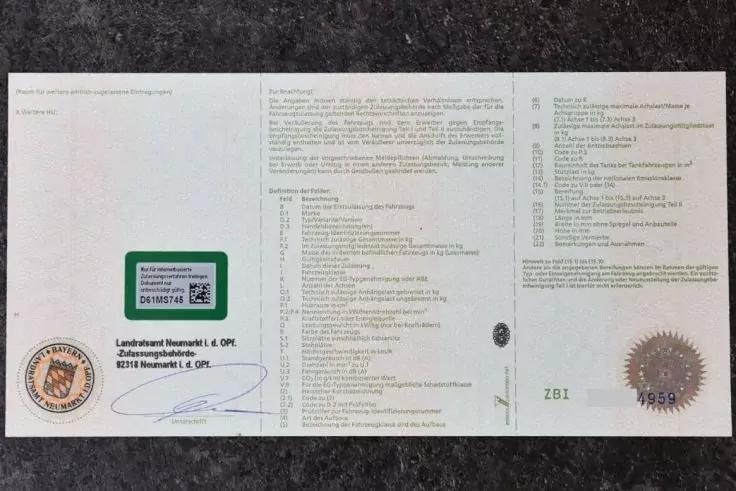 Wei&szlig;es, quer liegendes Dokument mit drei Spalten gedrucktem Text; links ein gr&uuml;ner Aufkleber mit QR‑Code, links unten ein runder blauer Stempel und eine handschriftliche Unterschrift, rechts unten ein goldener Pr&auml;gestempel; das Dokument liegt auf ein