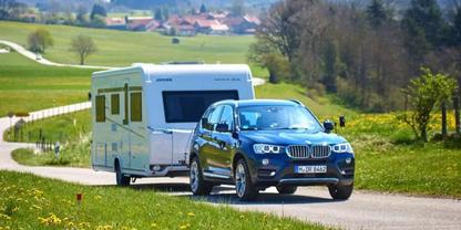 Dunkelblaues Auto zieht wei&szlig;en Wohnwagen auf einer Landstra&szlig;e, gr&uuml;ne Wiesen, H&auml;user und B&auml;ume im Hintergrund