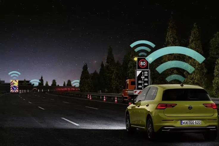 N&auml;chtliche Autobahnbaustelle, gr&uuml;ner Pkw von hinten, Fahrbahn mit Leitkegeln und Baustellenfahrzeugen, beleuchtete Schilder, mehrere stilisierte Wi‑Fi‑Symbole &uuml;ber Fahrzeugen und Schildern, B&auml;ume und dunkler Himmel