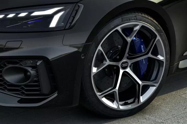 Nahaufnahme des vorderen Rads und Scheinwerfers eines dunklen Autos mit silberner Mehrspeichenfelge, blauem Bremssattel, Reifen und Lufteinlass