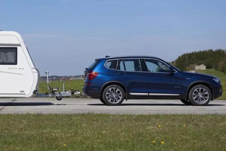 Blauer SUV zieht wei&szlig;en Wohnwagen auf einer Landstra&szlig;e; Wiese im Vordergrund und B&auml;ume am Horizont unter klarem Himmel