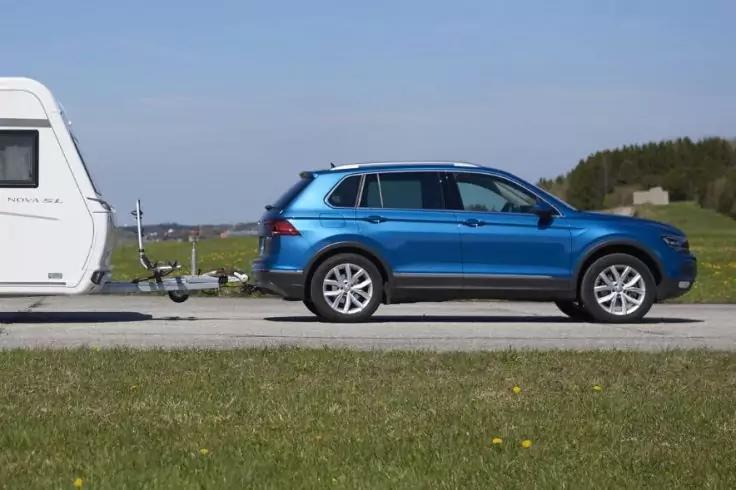 Blauer SUV mit angeh&auml;ngtem wei&szlig;em Wohnwagen auf einer Landstra&szlig;e, Gras im Vordergrund, B&auml;ume im Hintergrund, klarer Himmel